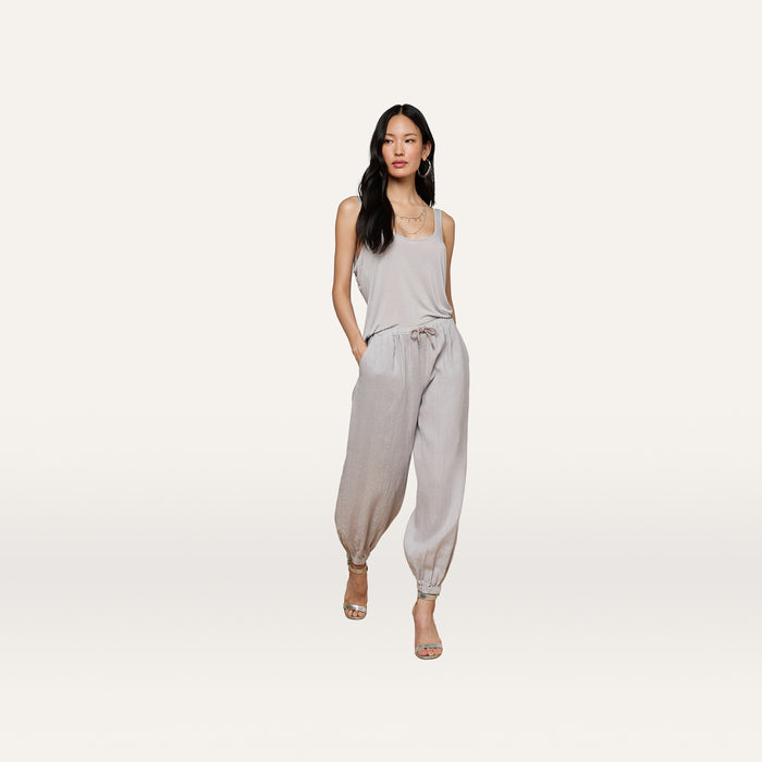 A woman stands against a plain white background, hands in pockets, wearing the Mariko Linen Jogger with a light beige sleeveless top, hoop earrings, and open-toe heels.