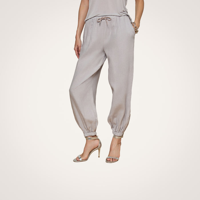 A person is shown from the waist down, modeling the Mariko Linen Jogger in light gray—featuring elastic cuffs and a drawstring waist—paired with a matching top and high-heeled sandals, set against a plain white background.