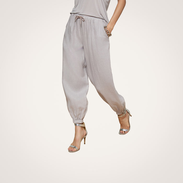 Model showcases the Mariko Linen Jogger in light gray, paired with a matching top and styled with silver high-heeled sandals. Posing with hands in pockets against a plain white background, the image highlights only the lower half of the outfit.