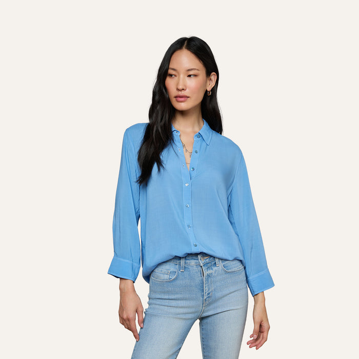 A woman with long black hair wears the Marlene Blouse, a relaxed fit, lightweight button-down shirt in light blue, paired with matching light blue jeans. She stands against a plain off-white background, looking slightly to her left with a relaxed expression.