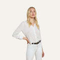 A woman with long blonde hair stands with one hand on her hip against a plain light background, wearing the Marlene Blouse—a relaxed fit white button-down shirt—paired with white pants and a black belt.