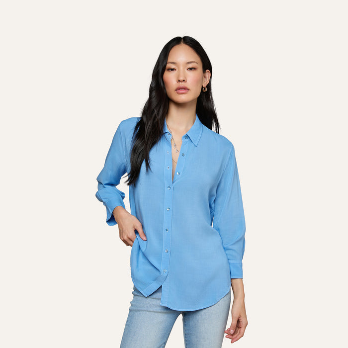 A woman with long black hair wears the Marlene Blouse—a relaxed fit, lightweight button-down shirt—paired with light jeans, standing with one hand in her pocket against an off-white background for effortless everyday polish.