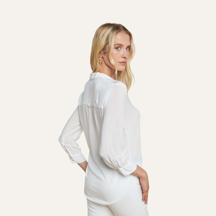 A woman with long blonde hair wears the Marlene Blouse and white pants, standing sideways and looking over her shoulder against a plain light background.