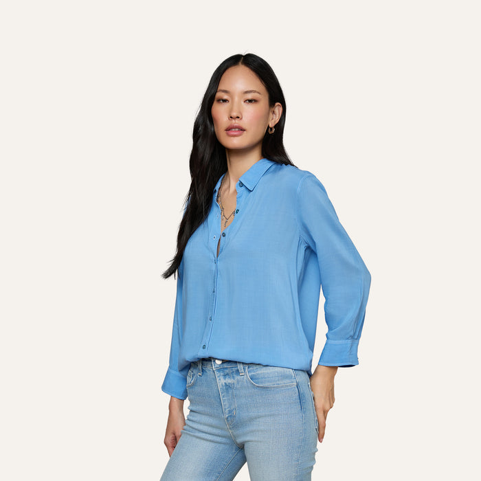 A woman with long black hair wears the Marlene Blouse—a lightweight, relaxed-fit button-down in light blue with rolled sleeves—paired with light blue jeans, standing against a plain background and embodying everyday polish.
