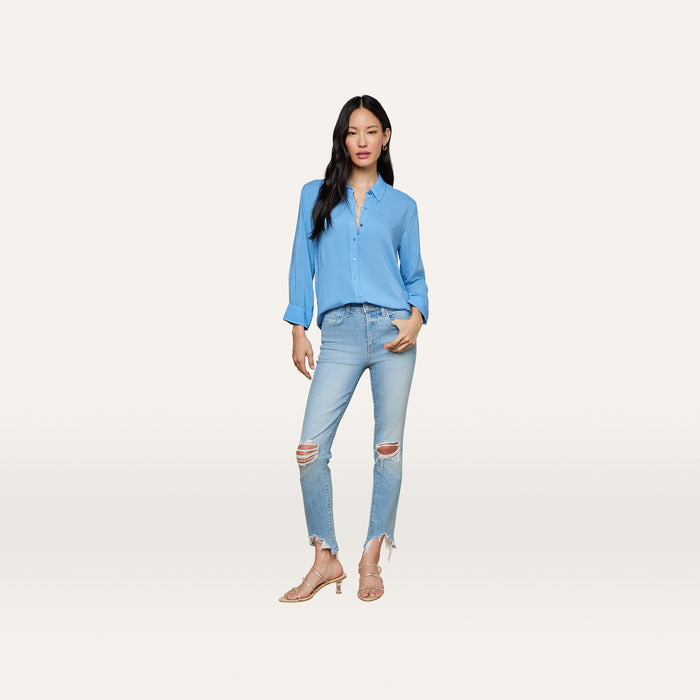 A woman wears the Marlene Blouse, a lightweight light blue button-down shirt, styled with ripped light-wash jeans and beige heels. She stands against a plain white background, facing forward with one hand in her pocket for an effortlessly relaxed fit and everyday polish.