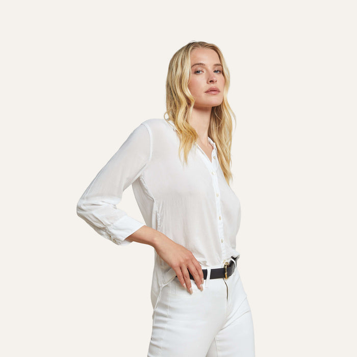 A woman with long blonde hair wears the Marlene Blouse—a classic white button-down shirt—paired with white pants and a black belt, standing against a plain light background and looking slightly to the side with a neutral expression.