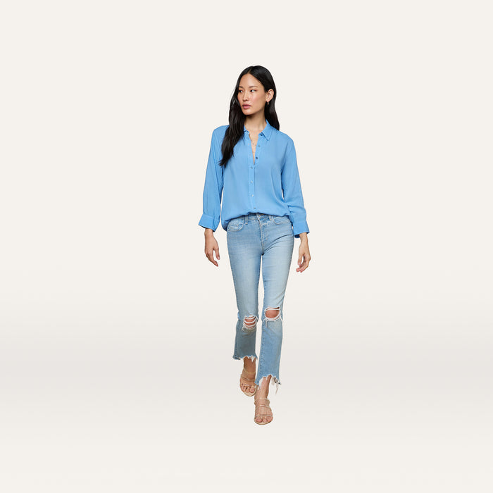 A woman wearing the Marlene Blouse, a relaxed-fit, lightweight button-down in light blue, pairs it with ripped light-wash jeans and beige sandals while standing against a plain white background for an effortless touch of everyday polish.