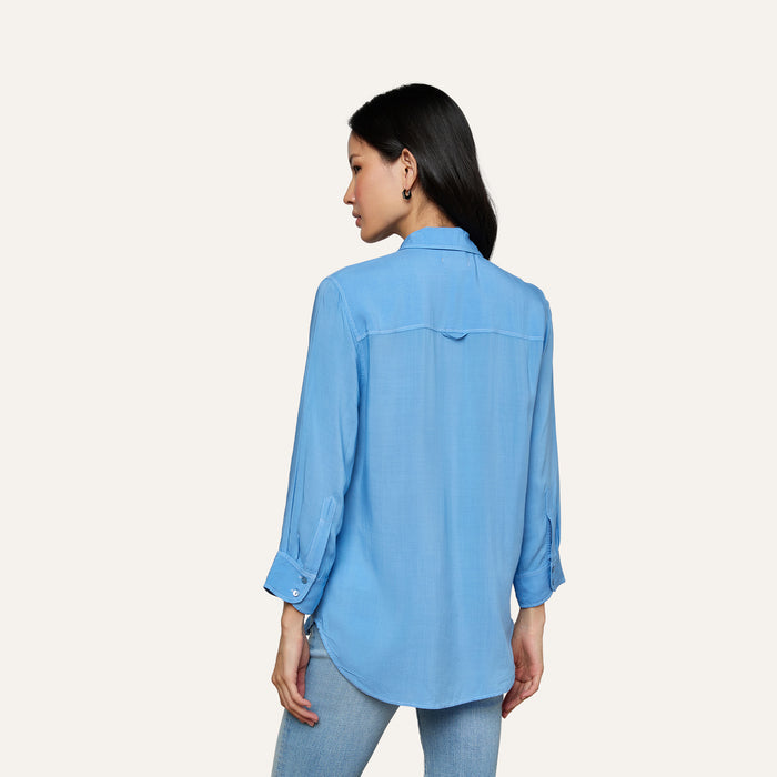 A woman stands facing away, wearing the Marlene Blouse—a lightweight button-down with rolled-up sleeves—paired with light blue jeans against a plain, light background, embodying everyday polish with its relaxed fit.