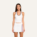 A woman stands facing forward against a plain light background, wearing the Martina Racerback Tennis Tank in white with a built-in bra and a matching skirt featuring red and blue side stripes.