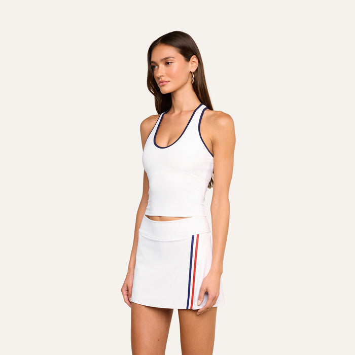 A woman wearing the Martina Racerback Tennis Tank, a sleeveless white tennis top, paired with a white skirt featuring red and blue side stripes, stands against a plain light background.