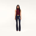 Wearing sunglasses, a sleeveless maroon turtleneck, pointed maroon shoes, and the Marty Coated Flare Jean, a woman stands against a plain white background.