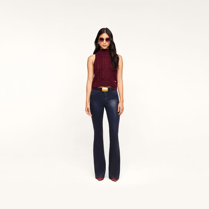 Wearing sunglasses, a sleeveless maroon turtleneck, pointed maroon shoes, and the Marty Coated Flare Jean, a woman stands against a plain white background.