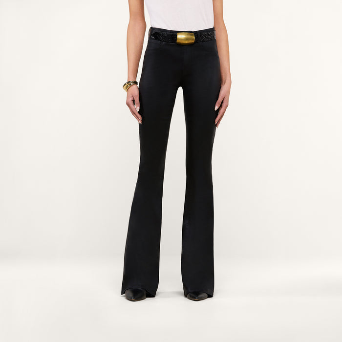 Wearing the Marty Coated Flare Jean in black, a person pairs the flared pants with a white sleeveless top, black shoes, a chunky belt featuring a gold buckle, and gold bracelets while standing against a plain white background.