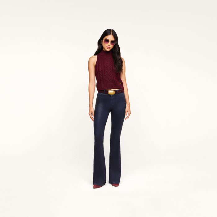 A woman wearing sunglasses, a sleeveless burgundy turtleneck sweater, a brown belt with a large buckle, pointed burgundy shoes, and the Marty Coated Flare Jean stands against a plain white background.