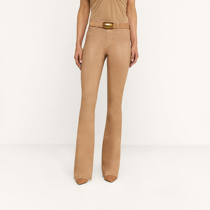 A person wearing the Marty Coated Flare Jean in tan stands against a plain white background. The outfit is styled with a matching belt featuring a large buckle and tan pointed shoes. The person’s upper body is not fully visible.