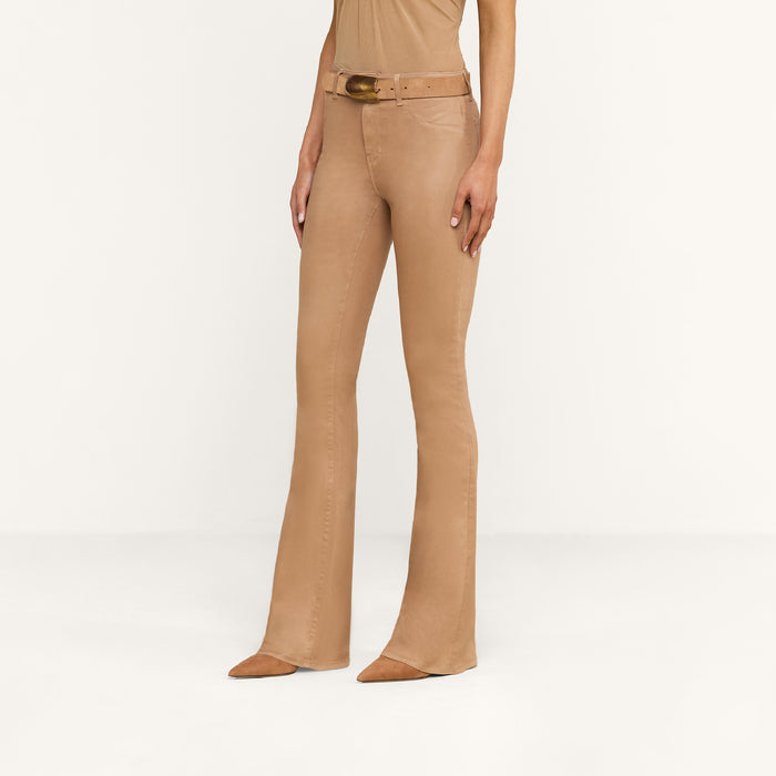 Wearing the Marty Coated Flare Jean—a pair of tan flare jeans crafted from a leather alternative—a person stands against a plain white background, styled with a matching top, wide belt, and pointed-toe shoes.