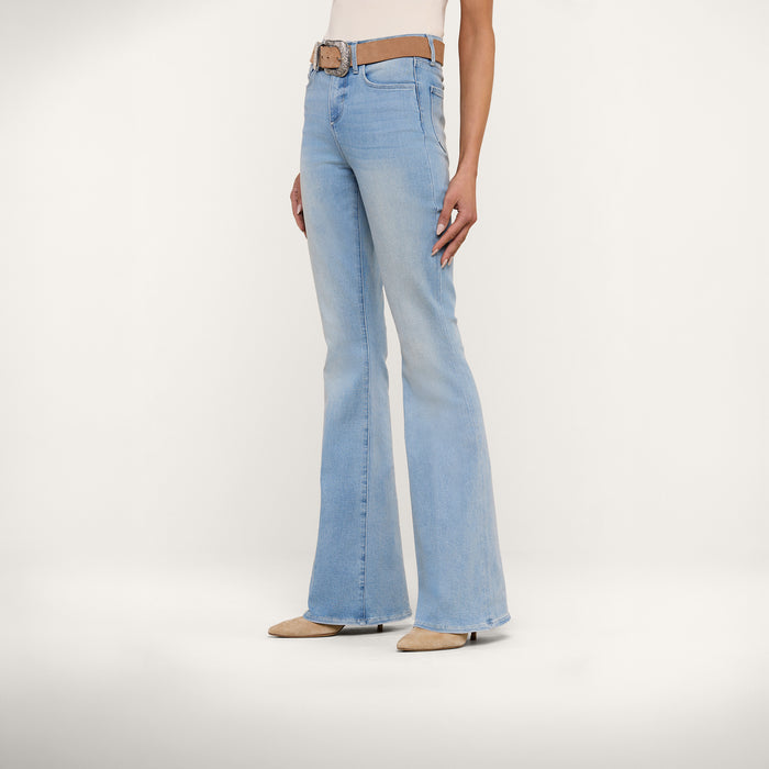 A person wearing the Marty Flare Jean in light blue, styled with a brown belt and beige pointed-toe shoes, stands against a plain white background. The upper body is mostly out of frame.