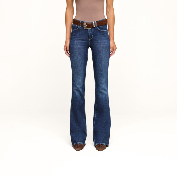 A person wearing a brown sleeveless top, the high-rise Marty Flare Jean crafted from stretch denim with a brown belt, and brown pointed-toe shoes stands against a plain white background.