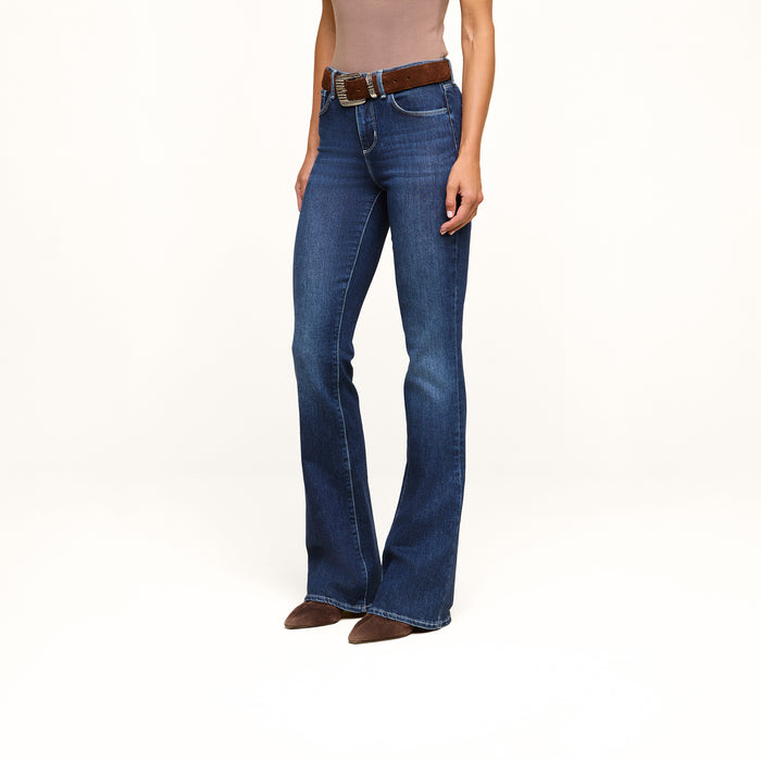 A person wearing the Marty Flare Jean in blue, styled with a brown belt and brown pointed-toe shoes, stands against a plain white background. Only the body from shoulders down is visible.