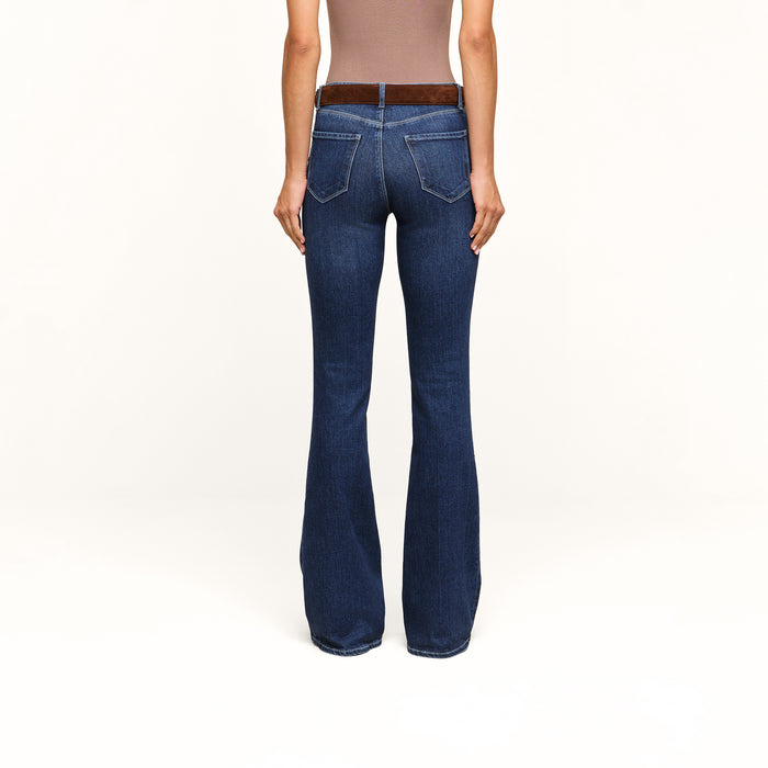 A person wearing a brown sleeveless top and the Marty Flare Jean in dark blue, viewed from the back, standing against a plain white background.