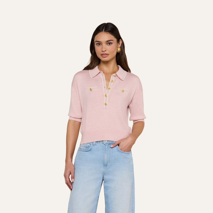 A woman stands against a plain white background wearing the Masaki Cropped Sweater—a light pink, featherweight knit polo top featuring gold buttons and front pockets—paired with light blue jeans.