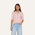 A woman stands against a plain white background wearing the Masaki Cropped Sweater—a light pink, featherweight knit polo top featuring gold buttons and front pockets—paired with light blue jeans.