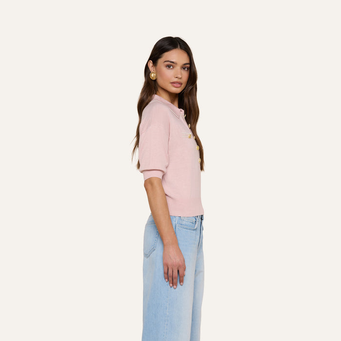 A woman with long, dark hair styled in loose waves stands in profile against a plain background, wearing the Masaki Cropped Sweater—a light pink featherweight knit polo top—paired with light blue high-waisted jeans and gold earrings.