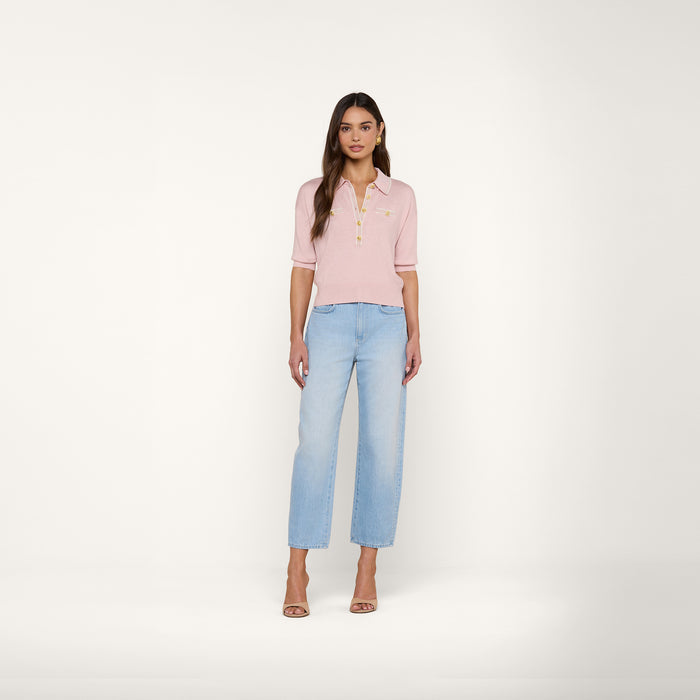 A woman stands against a plain white background, wearing the Masaki Cropped Sweater in light pink, paired with light blue straight-leg jeans and nude high-heeled sandals.