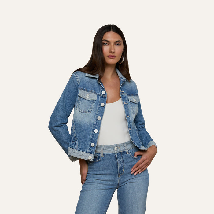 A woman stands against a plain light background with one hand on her hip, wearing the Maven Jean Jacket—a light-wash, three-quarter sleeve jean jacket—over a white top and blue jeans.