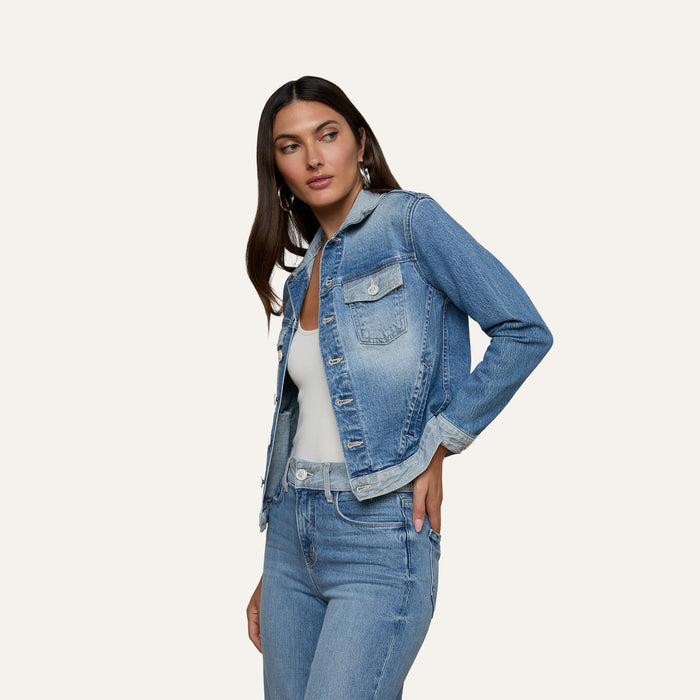 A woman with long dark hair wears the Maven Jean Jacket over a white top and matching blue jeans, standing against a plain light background, with one hand on her hip and looking slightly to the side.