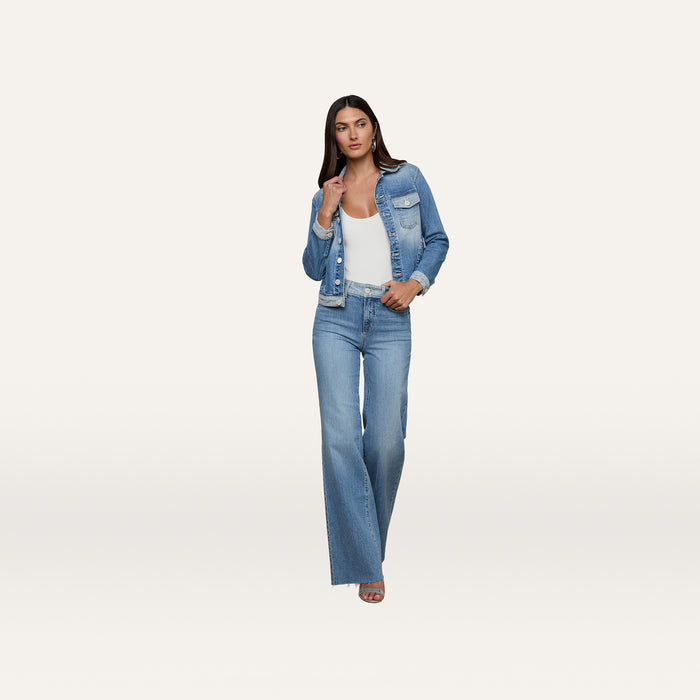 A woman wearing the Maven Jean Jacket with three-quarter sleeves, paired with wide-leg jeans over a white top, stands against a plain white background, looking forward with one hand touching her jacket.