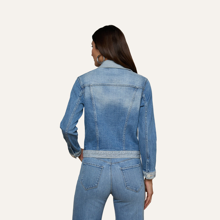 A woman with long dark hair is wearing the Maven Jean Jacket with three-quarter sleeves and matching stretch denim jeans. She is facing away from the camera, highlighting the back of her outfit against a simple light background.