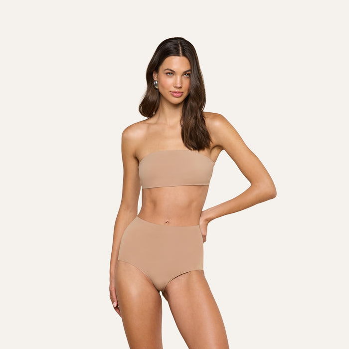 A woman stands against a plain light background with one hand on her hip and a relaxed expression, wearing the Mietta Knit Bandeau Top—a strapless, nude-colored bikini top paired with high-waisted stretch nylon bottoms.