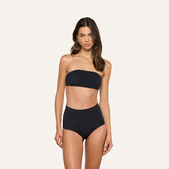A woman stands against a plain light background, wearing a black Mietta Knit Bandeau Top and high-waisted bikini bottoms. She has long brown hair and a neutral expression.