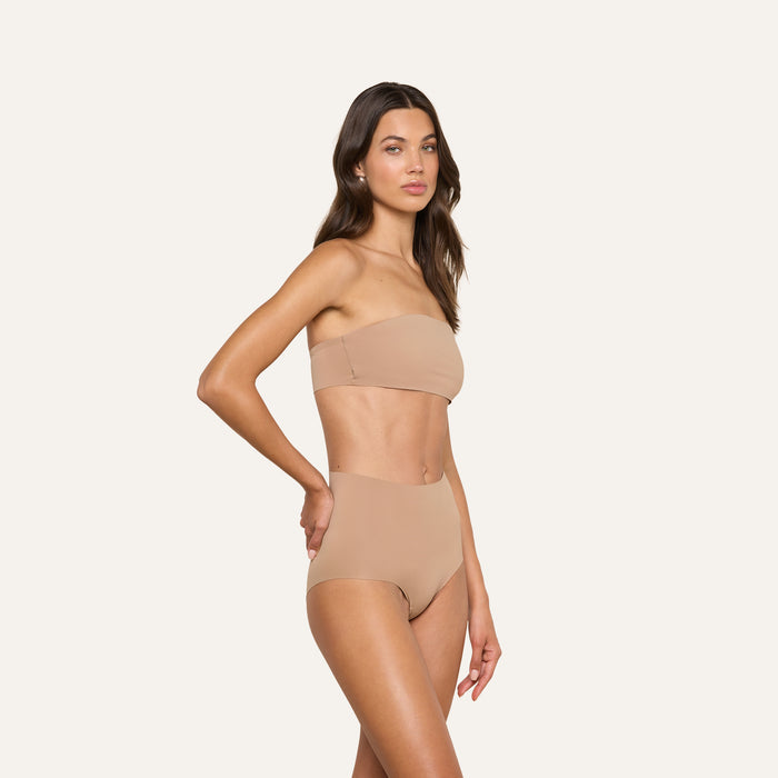 Wearing the Mietta Knit Bandeau Top, a woman poses against a plain white background in a strapless nude bikini top and high-waisted bottoms crafted from stretch nylon for a supportive fit.