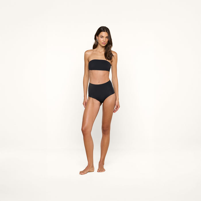 A woman stands against a plain white background, wearing the supportive Mietta Knit Bandeau Top in black paired with high-waisted bikini bottoms.