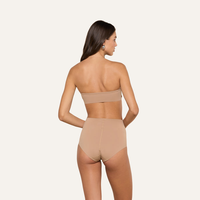 A woman stands facing away, wearing the Mietta Knit Bandeau Top in a strapless beige design, paired with high-waisted underwear. The set is crafted from stretch nylon for a supportive fit, set against a plain, light-colored background.