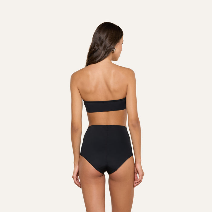 A woman with long brown hair stands with her back to the camera against a plain, light background, wearing the black Mietta Knit Bandeau Top paired with high-waisted bottoms.