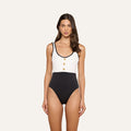 The model is wearing the Mila One-Piece Swimsuit, featuring a white top and black bottom with thin black straps and three gold buttons down the front. This high-leg design evokes French glamour as she stands facing forward against a plain, light background.