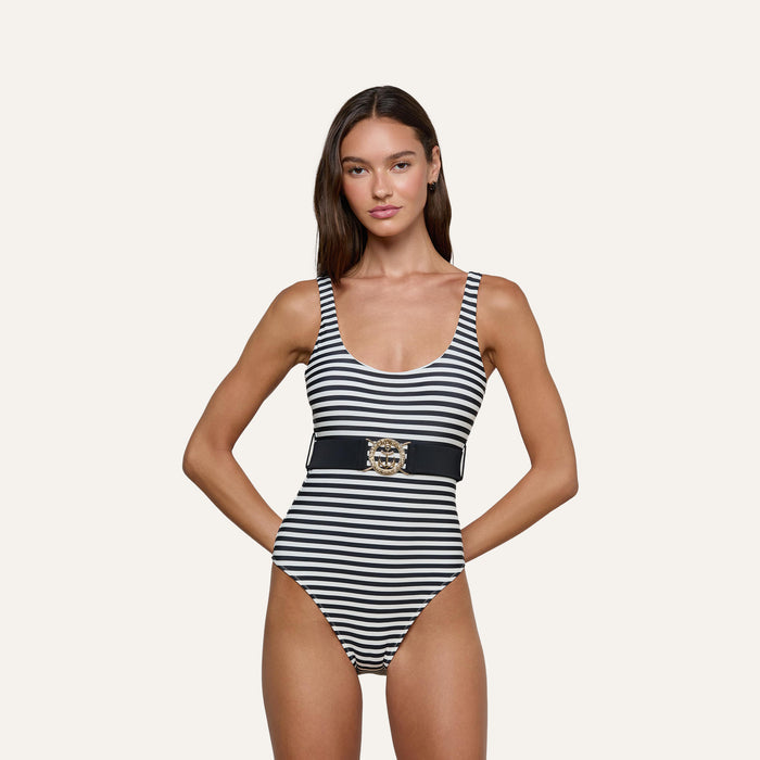 A woman stands with hands on hips against a plain background, wearing the Mila One-Piece Swimsuit—a black and white striped style featuring a wide belt with a decorative gold buckle and a scoop neckline.