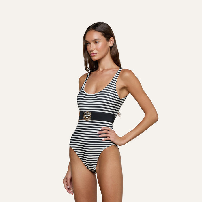 A woman stands with her hand on her hip against a plain background, wearing the Mila One-Piece Swimsuit: a black and white striped design featuring a scoop neckline and a wide black belt adorned with a decorative gold emblem.