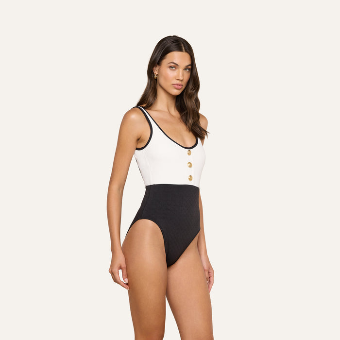 Model showcases the Mila One-Piece Swimsuit, a sleeveless black and white design adorned with gold buttons on the front. This high-leg swimwear style exudes subtle French glamour and is set against a plain beige background.