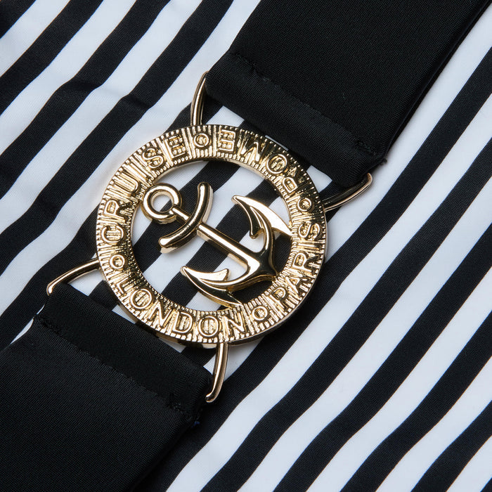 A close-up of a gold anchor belt buckle with engraved text, attached to a black strap, set against the bold black and white vertical stripes of the Mila One-Piece Swimsuit.