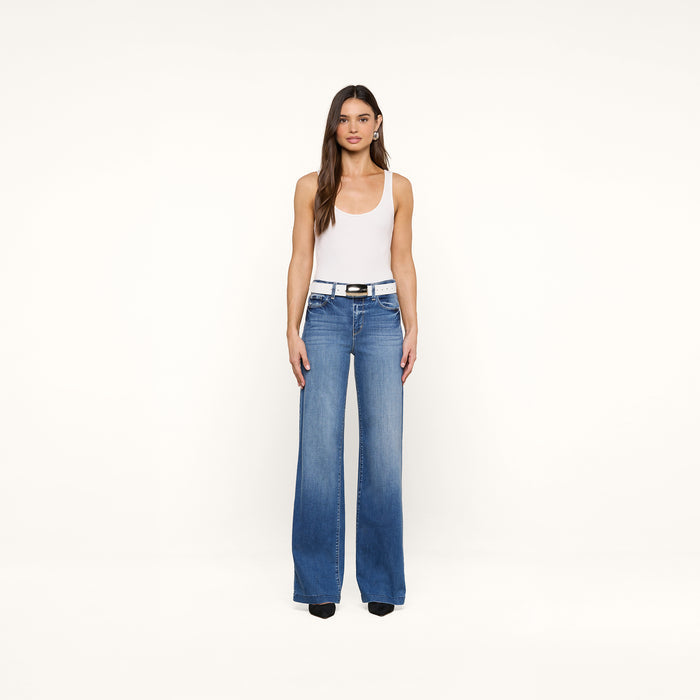 A woman with long brown hair wears a white tank top, blue high-waisted wide-leg jeans, black shoes, and the handcrafted Milou Belt, standing against a plain beige background.