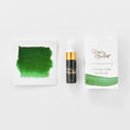 A mini ink sample bottle of New Forest Green fountain pen ink by Tom’s Studio, displayed alongside a swatch of the ink on textured paper and its matching packaging. The bottle is small, with a gold cap and a white pipette dropper, and features 'Tom’s Studio' in gold lettering. The ink swatch shows a rich, shimmering hue with a gradient from deep to lighter tones. The packaging, a small white card, has 'Tom’s Studio' embossed in gold at the top, with a brushstroke of the ink colour at the bottom.