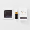 A mini ink sample bottle of Stellar black shimmer fountain pen ink by Tom’s Studio, displayed alongside a swatch of the ink on textured paper and its matching packaging. The bottle is small, with a gold cap and a white pipette dropper, and features 'Tom’s Studio' in gold lettering. The ink swatch shows a rich, shimmering hue with a gradient from deep to lighter tones. The packaging, a small white card, has 'Tom’s Studio' embossed in gold at the top, with a brushstroke of the ink colour at the bottom.