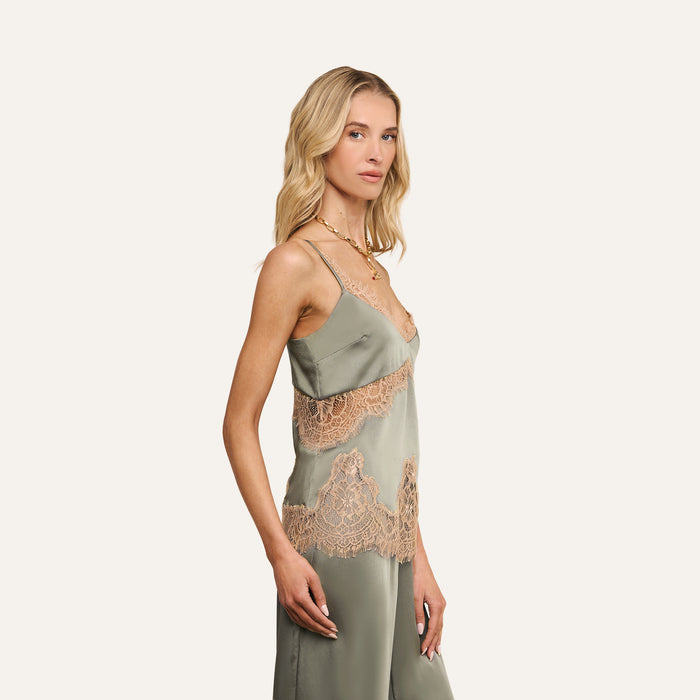 A woman with long blonde hair stands wearing the Mitsu Satin Lace Camisole, a sage green, lingerie-inspired top with beige lace accents, paired with matching pants. She looks toward the camera against a plain, light-colored background.