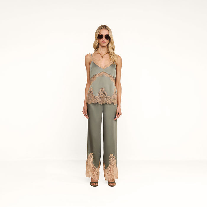 A woman stands facing forward in a minimal white studio, wearing sunglasses and the Mitsu Satin Lace Camisole in green paired with matching pants featuring beige lace trim.