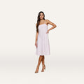 A woman stands against a plain white background wearing the Mitzy Linen Midi Dress, a sleeveless, light pink linen day dress with adjustable straps, paired with beige high-heeled sandals. Her hand rests on her hip as she looks at the camera.