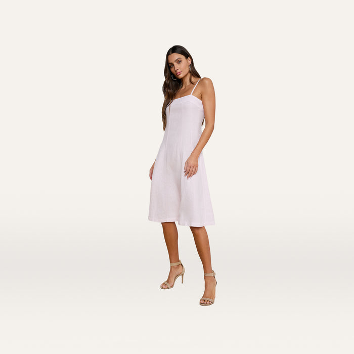 A woman stands against a plain white background wearing the Mitzy Linen Midi Dress in light pink, featuring adjustable straps. She pairs the dress with beige high-heeled sandals, her long brown hair down, and poses with one knee slightly bent.